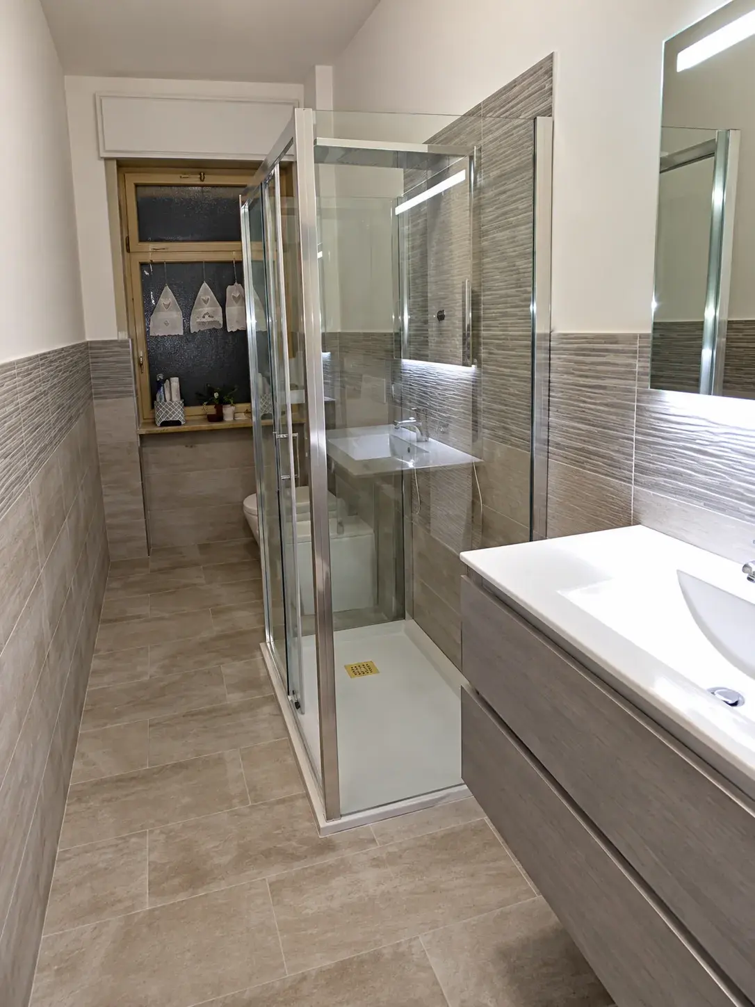 Bagno con box doccia in vetro, rivestimenti grigi e mobile lavabo
